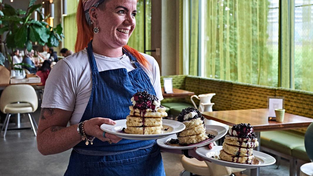 Woman in a blue apron holding plates of layered pancakes in a modern restaurant with large windows.